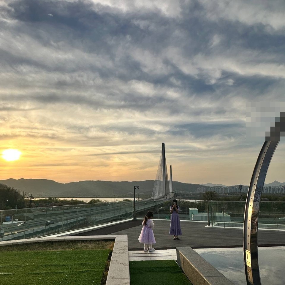 강동 아이파크 더 리버몰 야외 테라스에서 보라색 드레스를 입은 아이와 성인이 석양을 배경으로 서 있는 모습, 유리 난간과 잔디가 보임