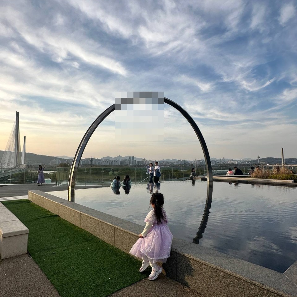 아이파크리버몰 야외 테라스의 원형 아치 조형물과 얕은 수반, 보라색 드레스 어린이가 물가에 앉아 있고 도시 전경이 배경